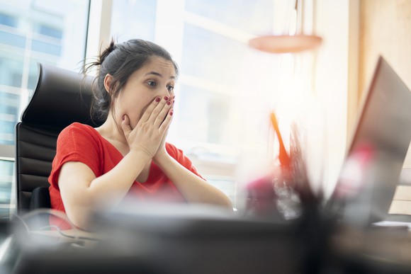A woman sitting at her computer with her hands over her mouth as if surprised.