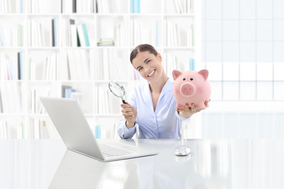 A woman with a piggy bank and a magnifying glass in front of her laptop.