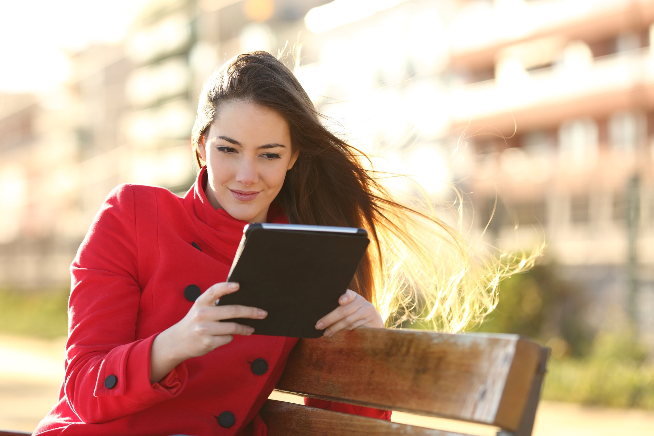 A smiling young woman in a red coat sits on a park bench, using a tablet computer.