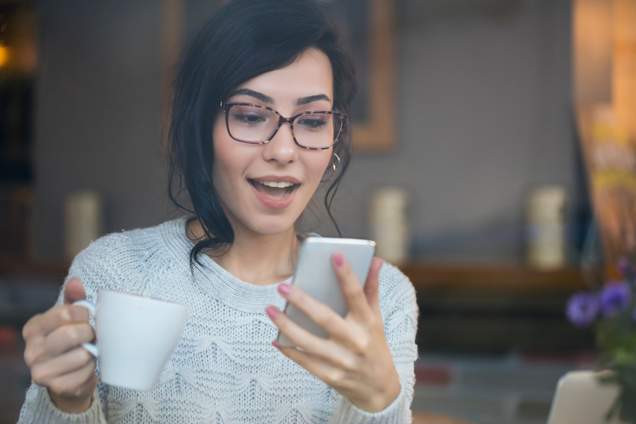 Woman looking excitedly at her smartphone while holding a mug.