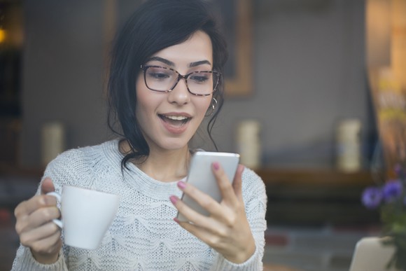 Woman looking excitedly at her smartphone while holding a mug.
