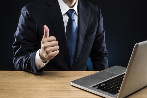 Man wearing coat and tie giving a thumbs up in front of a laptop