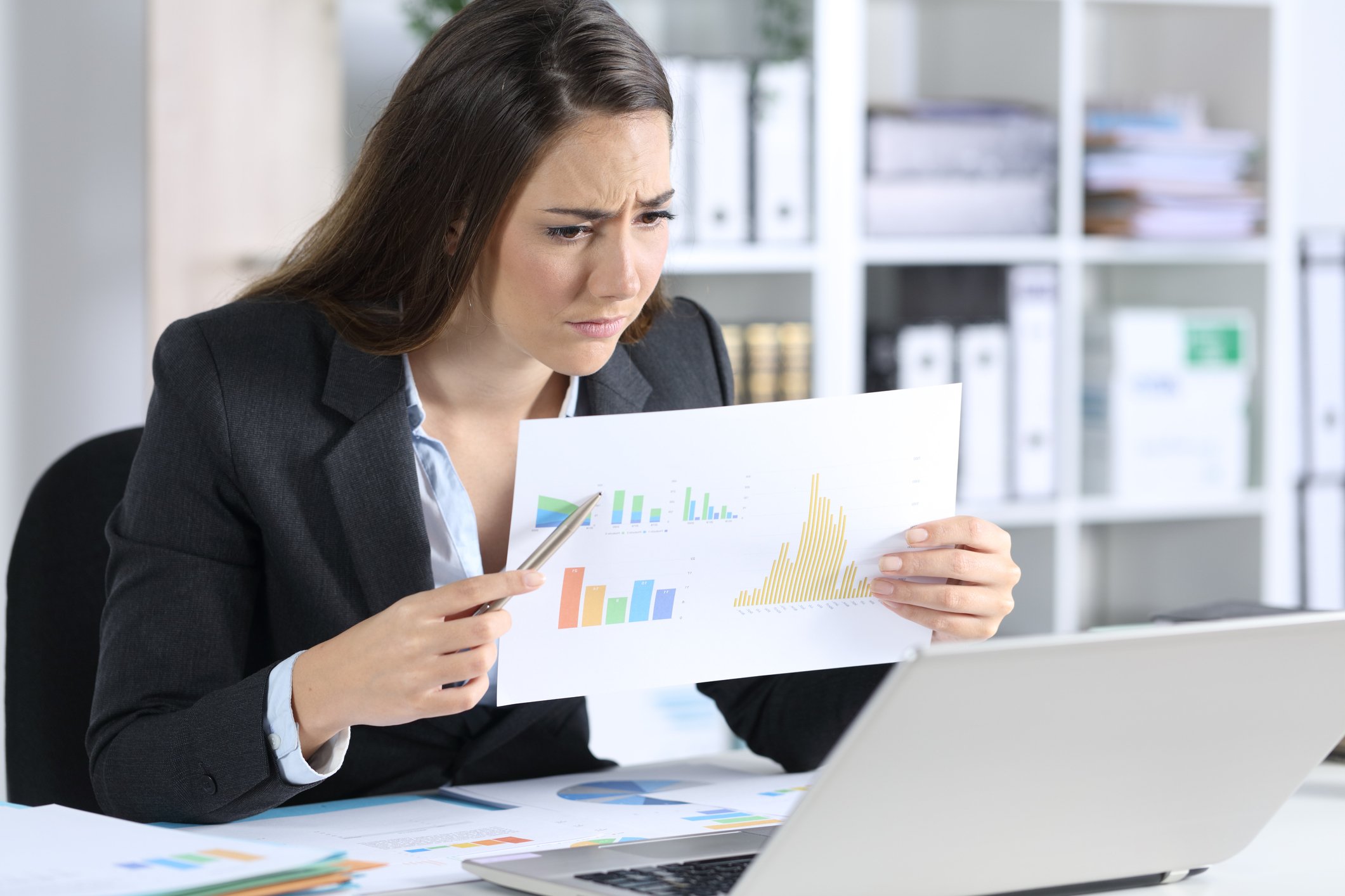 A young businesswoman frowns, holding up a paper full of charts to her laptop's webcam.