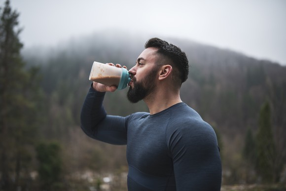 An outdoor fitness enthusiast drinking a protein shake in front of a misty, forested hill.