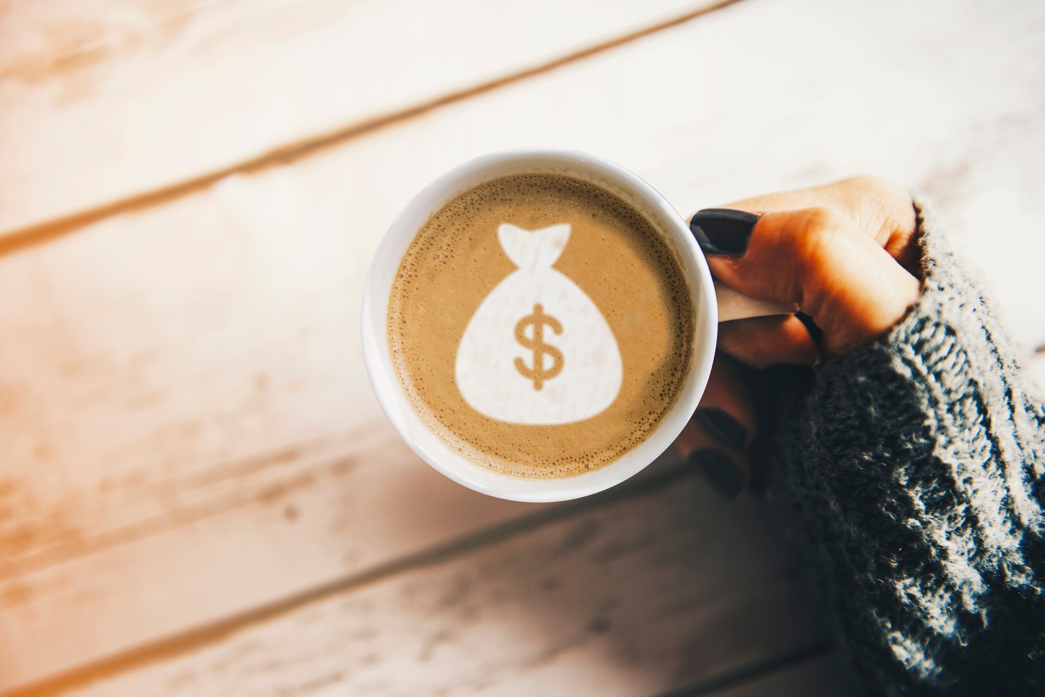 A woman's hand with black nail polish holding a coffee cup with a money bag symbol on the coffee surface.