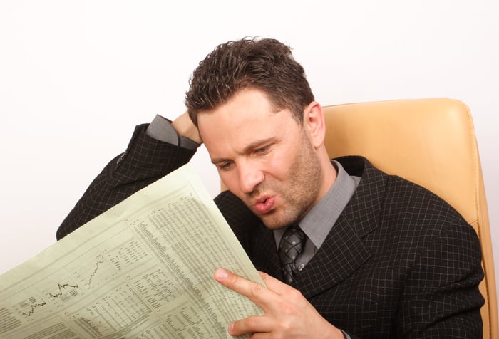 A surprised man reading a financial newspaper with visible stock quotes. 