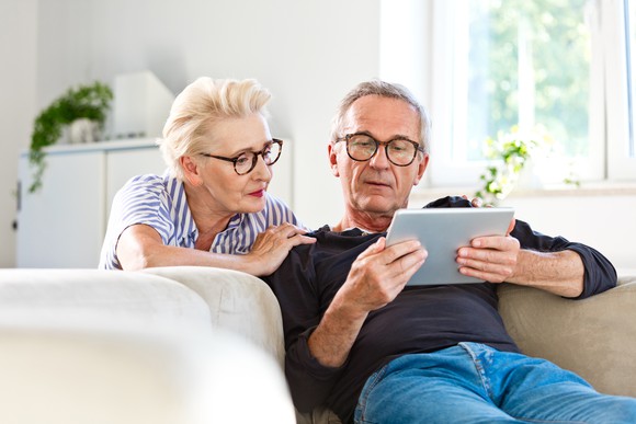 An elderly couple sitting on a couch looking at data on a tablet. 