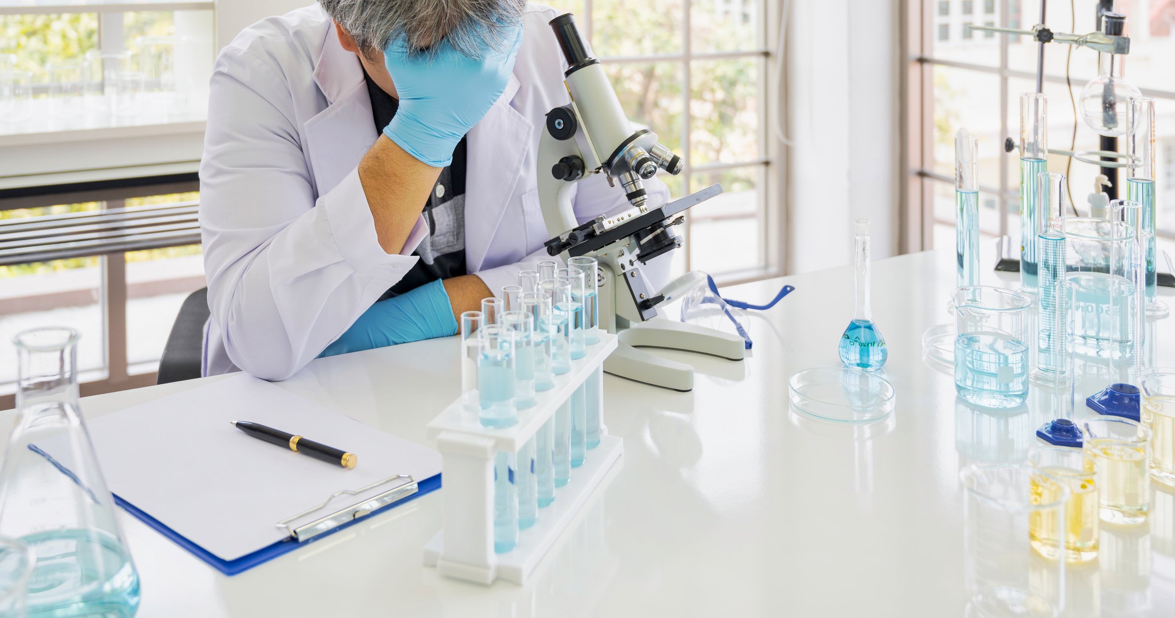 A scientist sits in front of a microscope near a rack of test tubes, face covered.