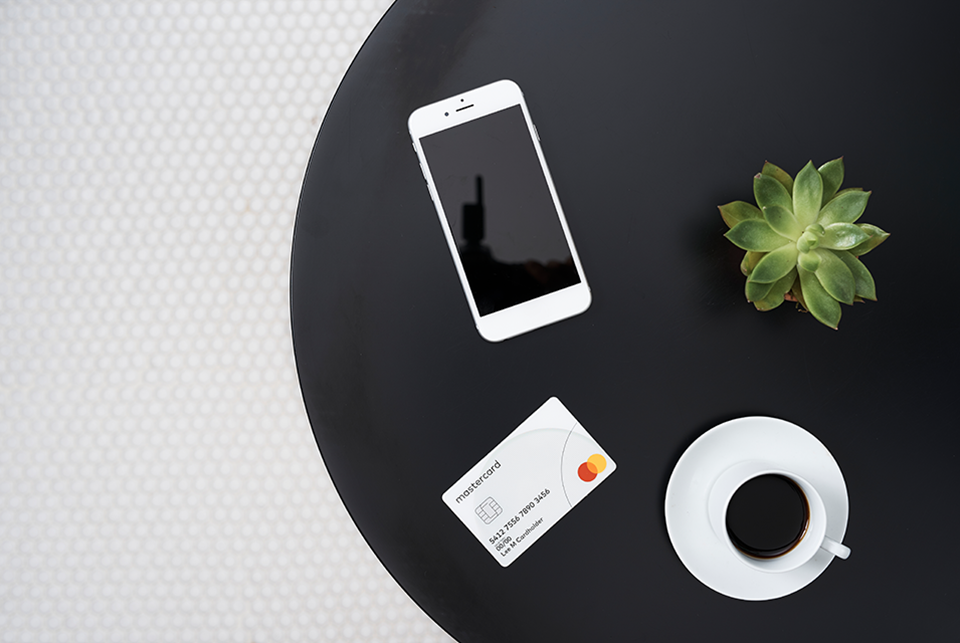 A Mastercard branded credit card sits on a table between a smartphone and a cup of coffee.