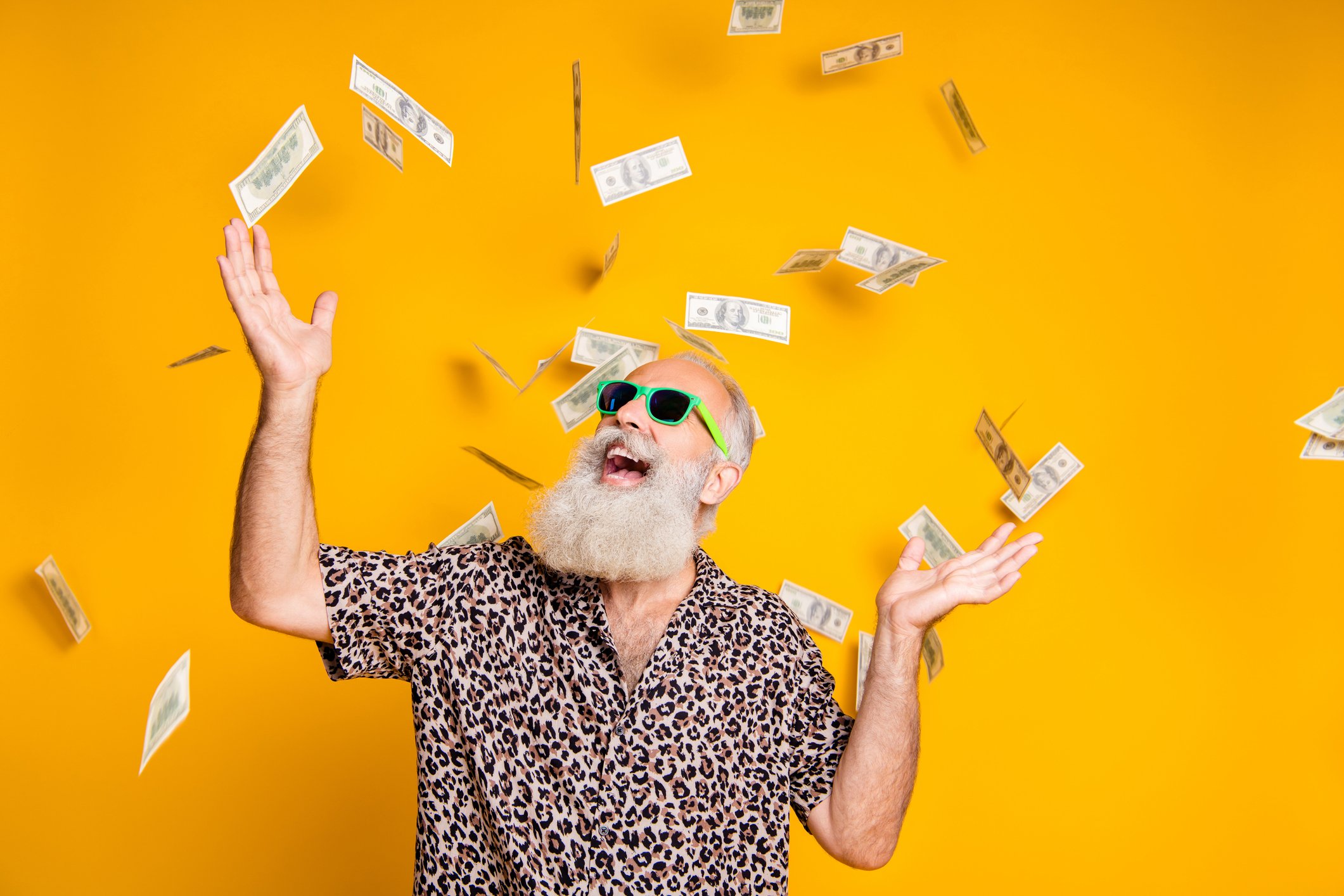 An elderly bearded man wears sunglasses while cash money rains down.