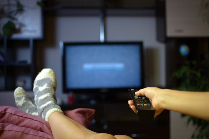 Pointing a connected TV remote at a television screen, with propped-up feet.