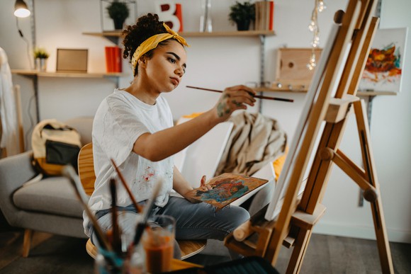 Seated young woman holding a paint brush while considering her easel-supported canvas.