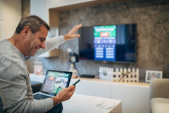 A man watching his stock portfolio on his phone with a laptop and TV monitor in front of him also showing stock market data