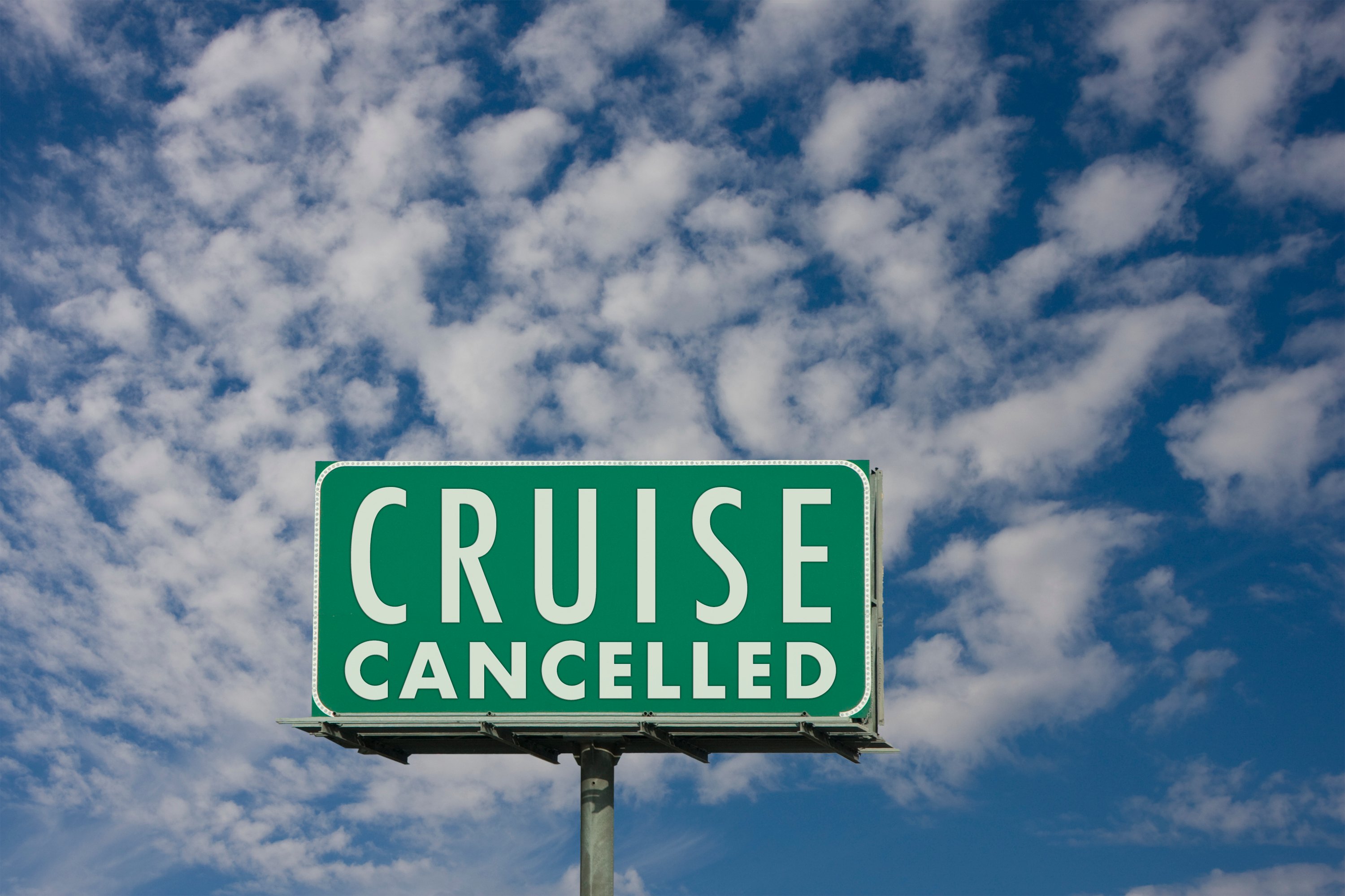 A green billboard with white lettering reading "Cruise Cancelled" against a blue sky with cirrocumulus clouds.