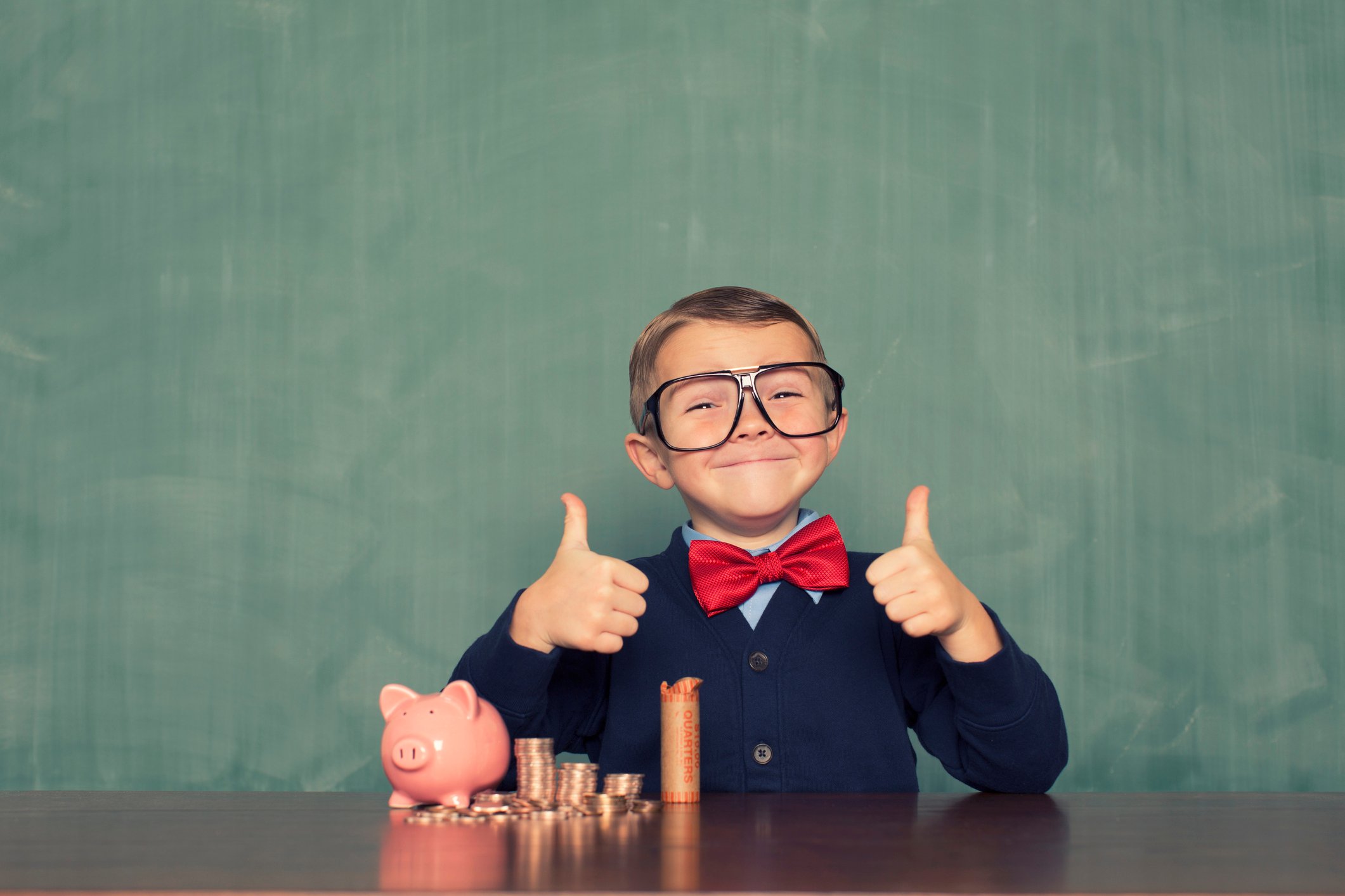 young kid wearing oversized glasses with coins and piggy bank out on the table and giving a thumbs up