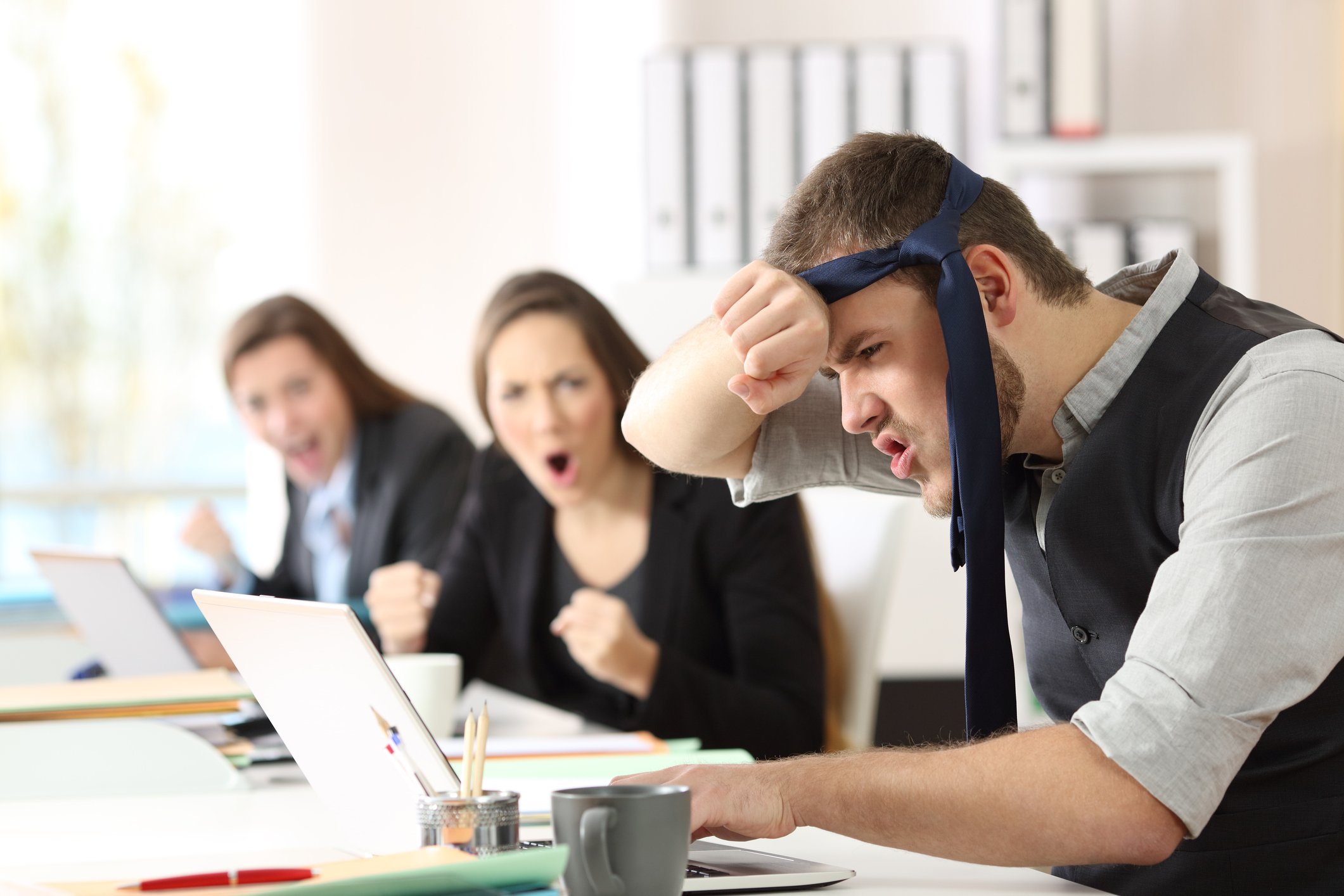 A young office worker wipes the sweat off his forehead, wearing his tie as a bandana. Two female colleagues cheer him on.