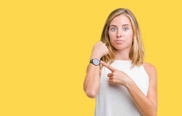 A woman looks at the camera and points to her wristwatch.
