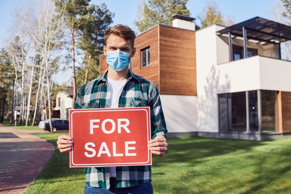 Young man wearing a health mask holds a "For Sale" sign in front of a house.