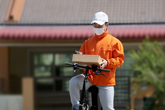 A deliveryman on a bike looks at a package in hishands wearing a mask. 
