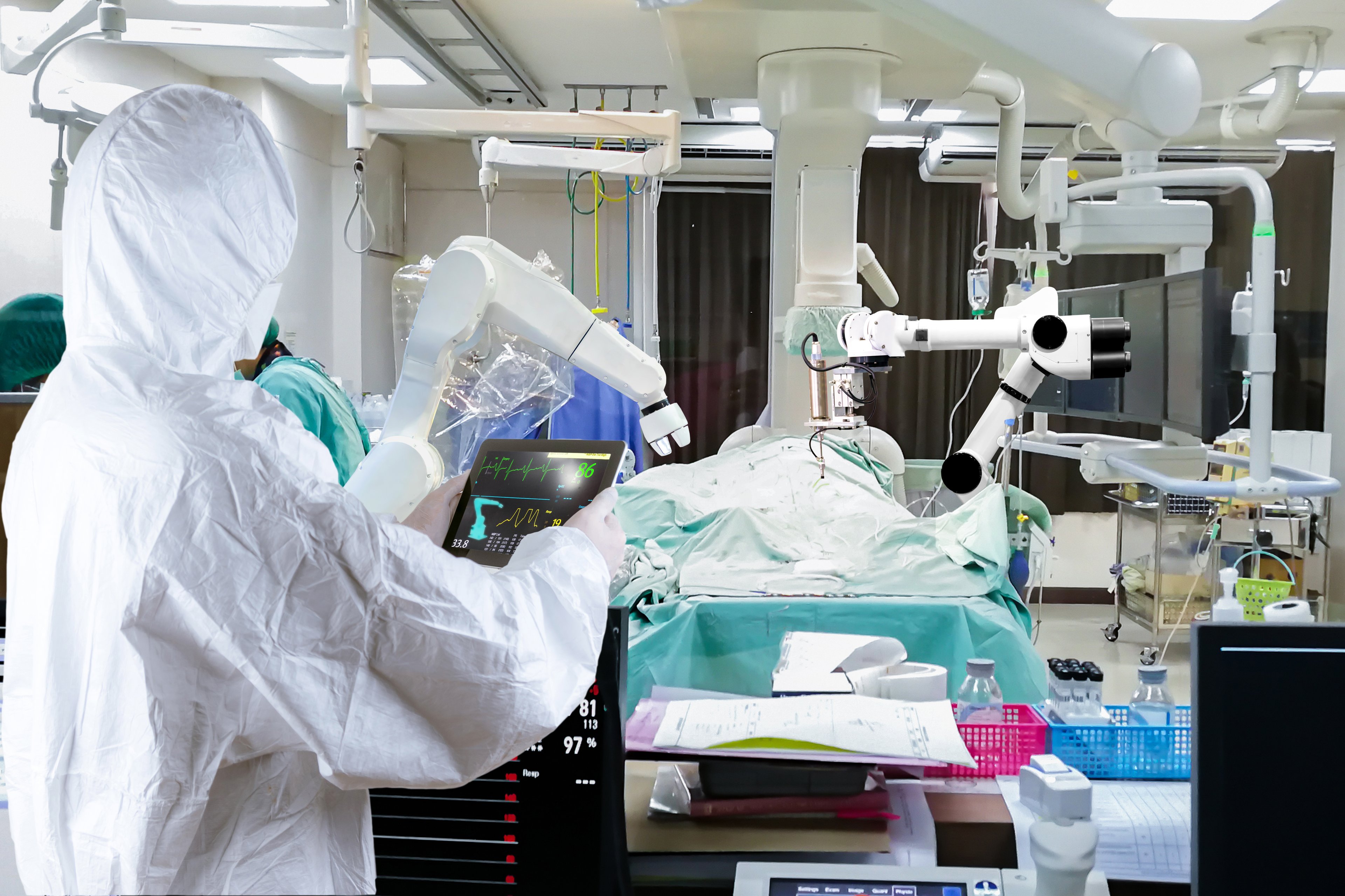 Surgeon wearing full protective gear holds a portable tablet which controls a remote-operated machine in the operating room.