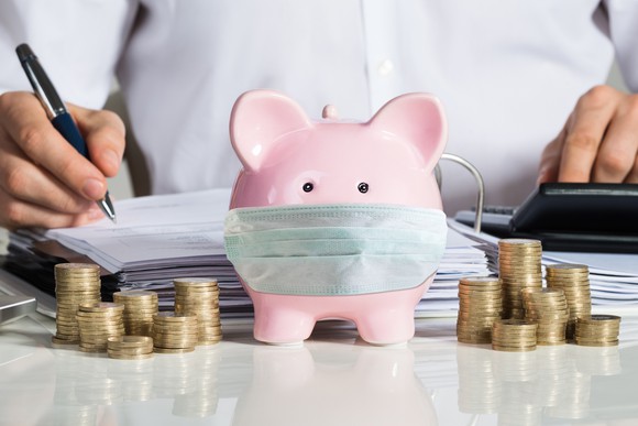Piggy bank wearing a face mask on a table surrounded by coins, documents, and a man sitting at the desk.
