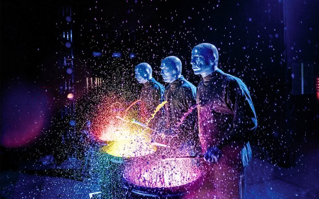 Three Blue Man Group performers playing drums in colored water. 