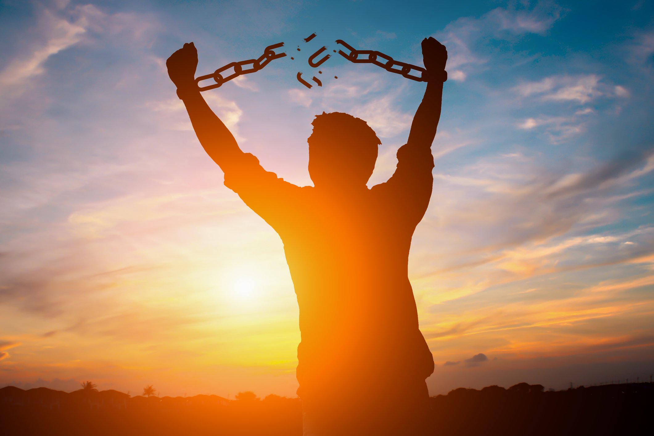 Silhouette of a man breaking his shackles against a colorful sunrise.
