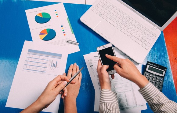 Two pairs of hands holding a phone and pen, surrounded by printed graphs, a calculator, and a laptop.