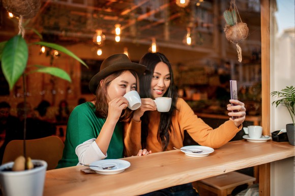 Two friends sip coffee at a coffeeshop and take a selfie. 