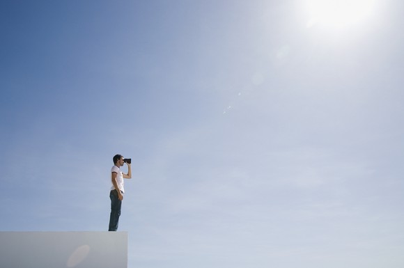 Man under blue skies looking through binoculars into the distance.