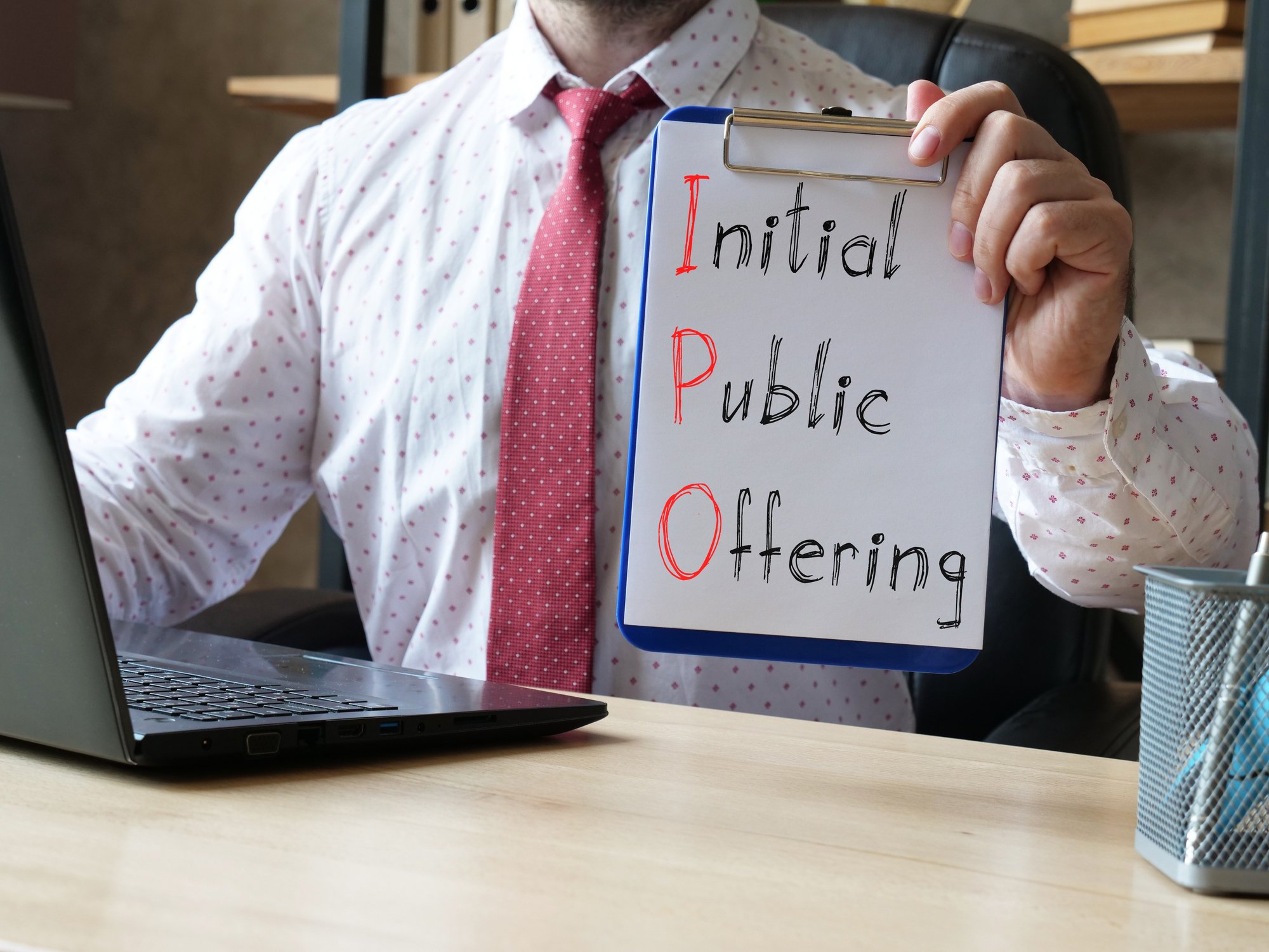 A man sitting at a desk in front of a computer holding a clipboard that reads "initial public offering" with the letters "IPO" highlighted.