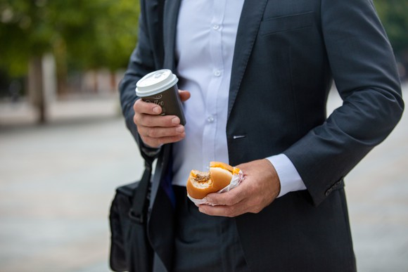 man standing with a coffee cup in right hand and burger in left hand
