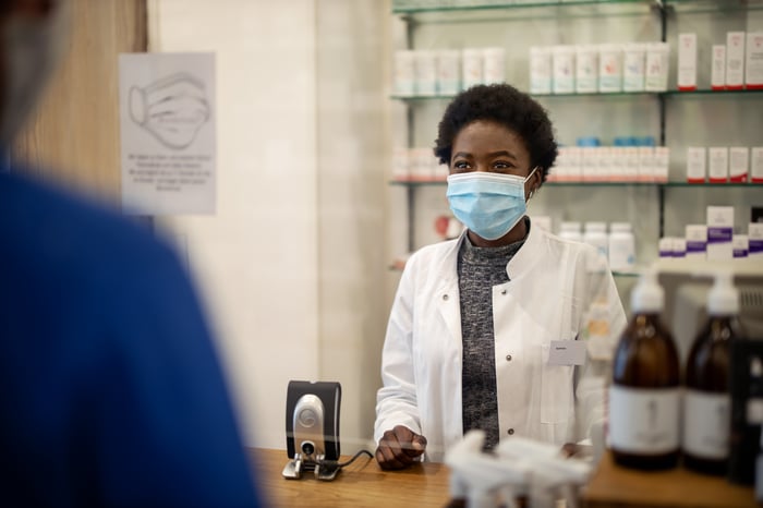 Female pharmacist wearing a mask and white coat stands behind a counter, in front of a wall of white pill bottles, greeting customer in blue shirt.