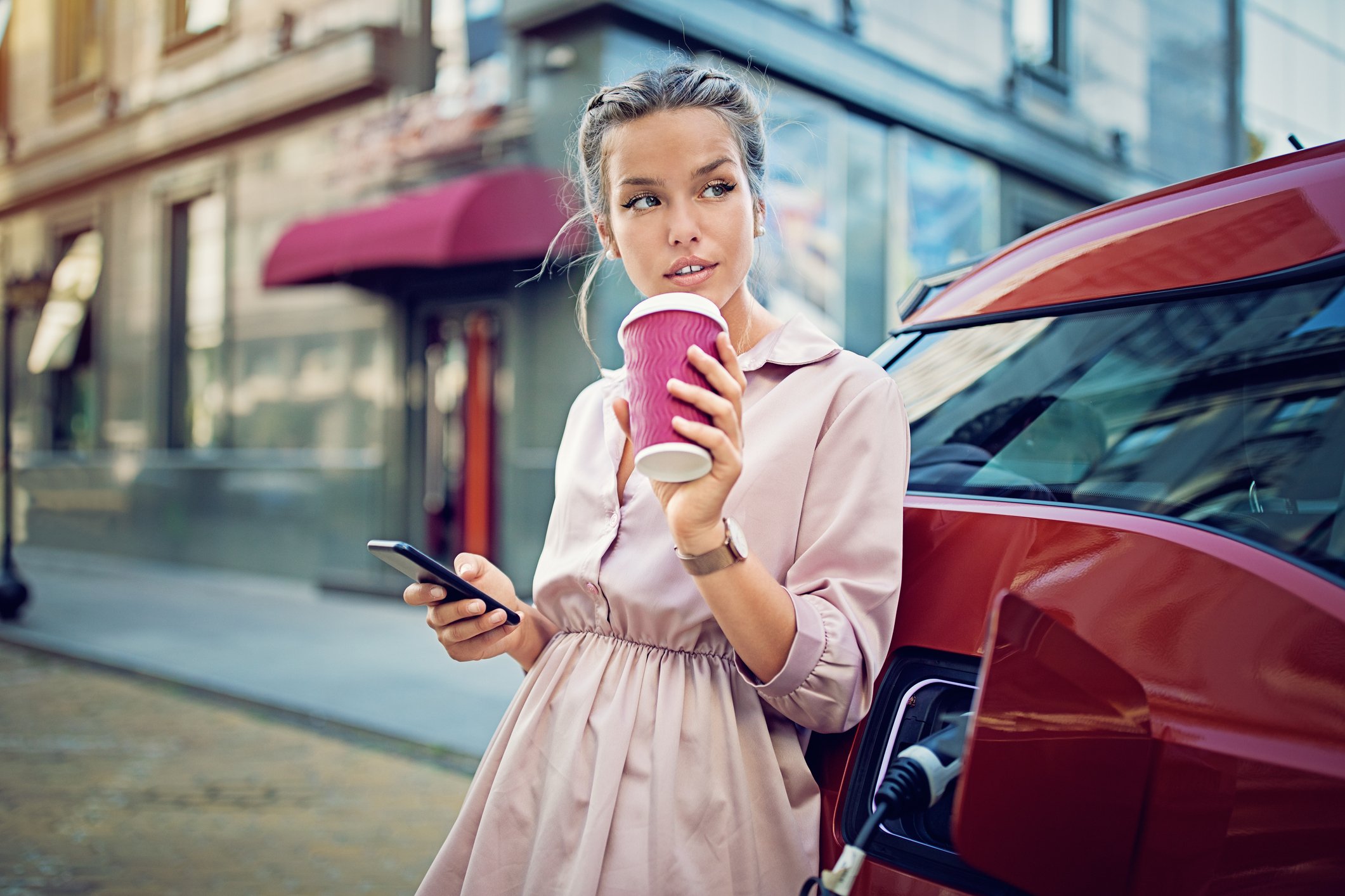 A woman drinking a beverage and looking at her phone leans against an electric vehicle while it charges.