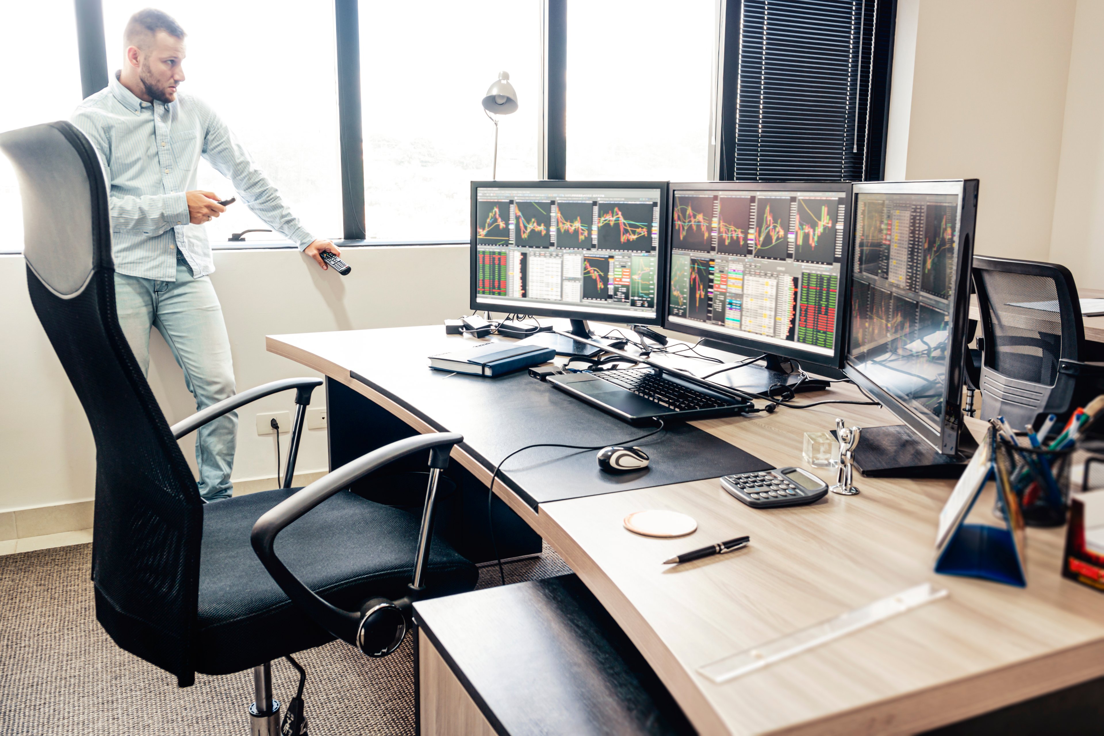 A person checks their cellphone while standing at the window of an office with a desk that has several monitors displaying charts. 