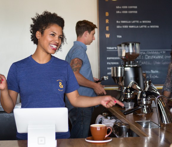 Barista smiling at customer from behind a coffee bar, using a Square terminal