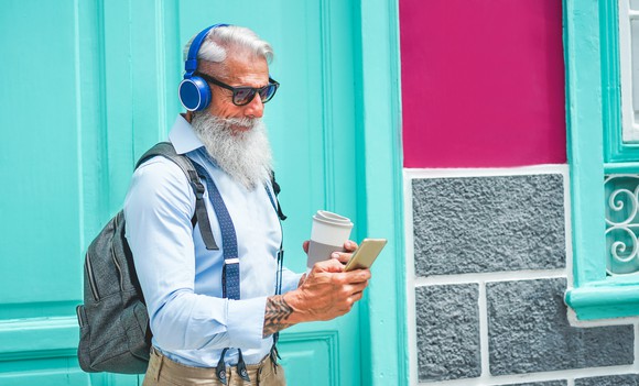 A trendy senior man walks through a downtown center holding a coffee and smartphone.