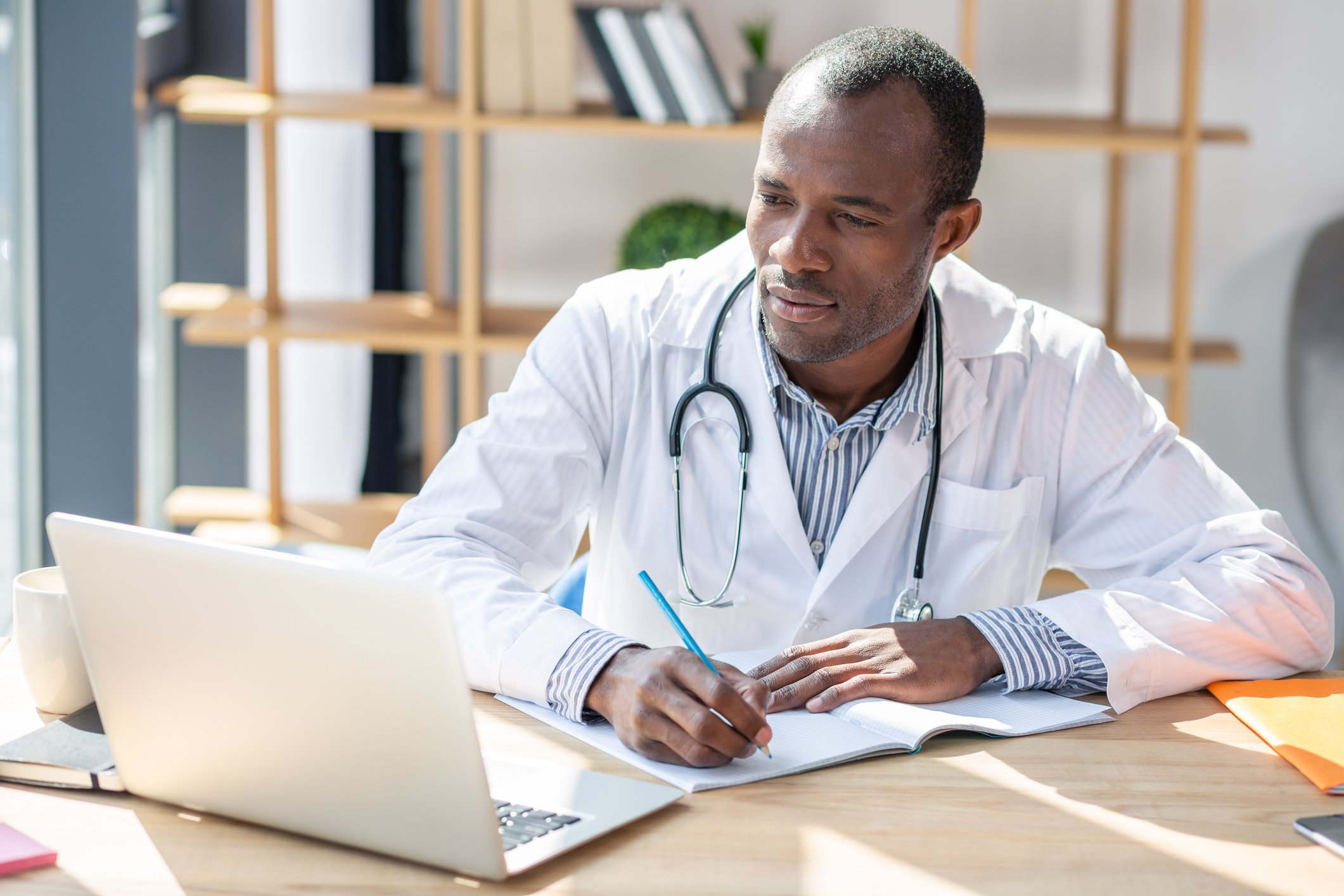 Male healthcare provider writing in notebook while looking at laptop.