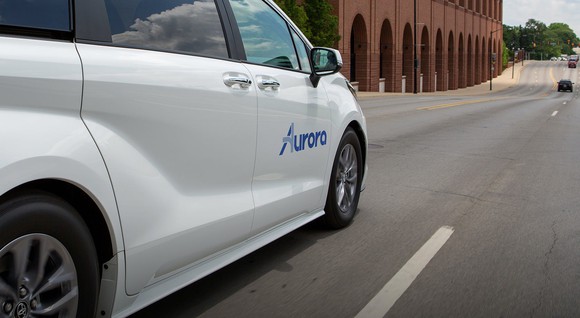 A white Toyota minivan with an Aurora logo on its door drives down a street.