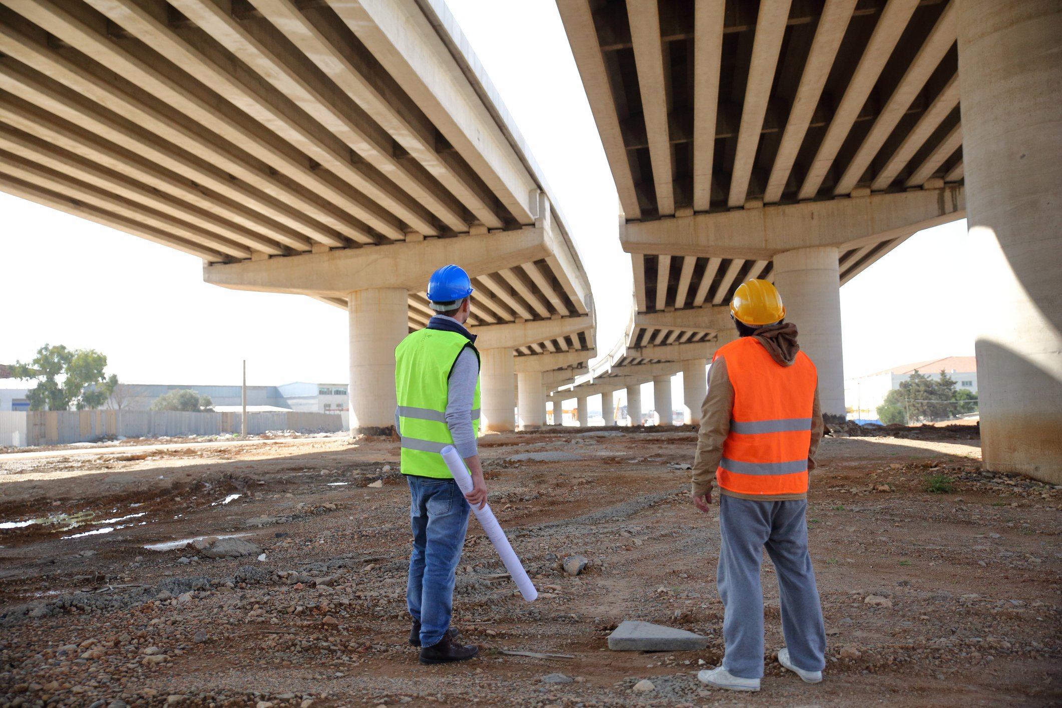 Two workers stand underneath a highway project.