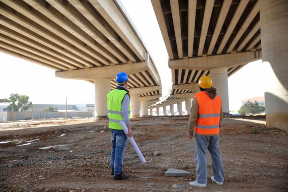 Two workers stand underneath a highway project.