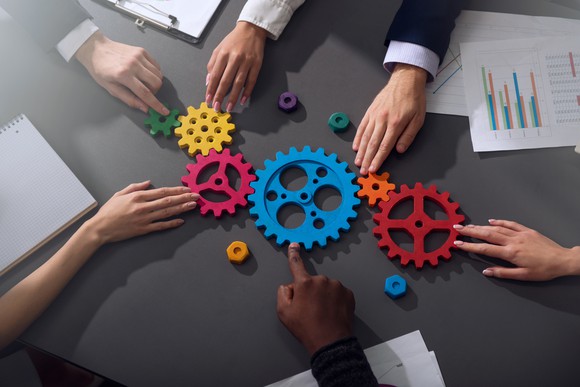 A set of colored cog wheels sitting on a table, with business people interacting with them.