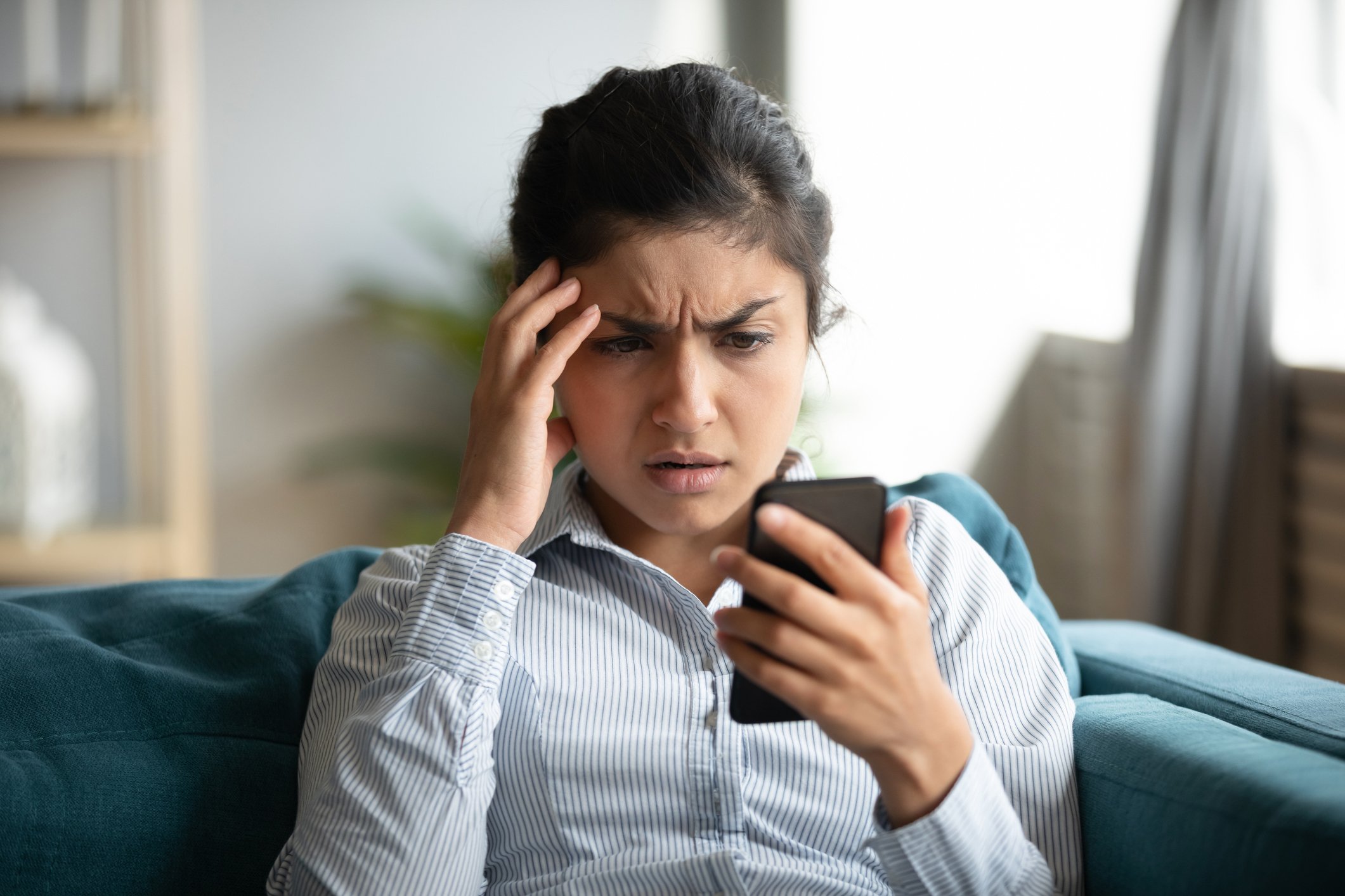 A frustrated woman, with her hand to her head, looks at her phone.