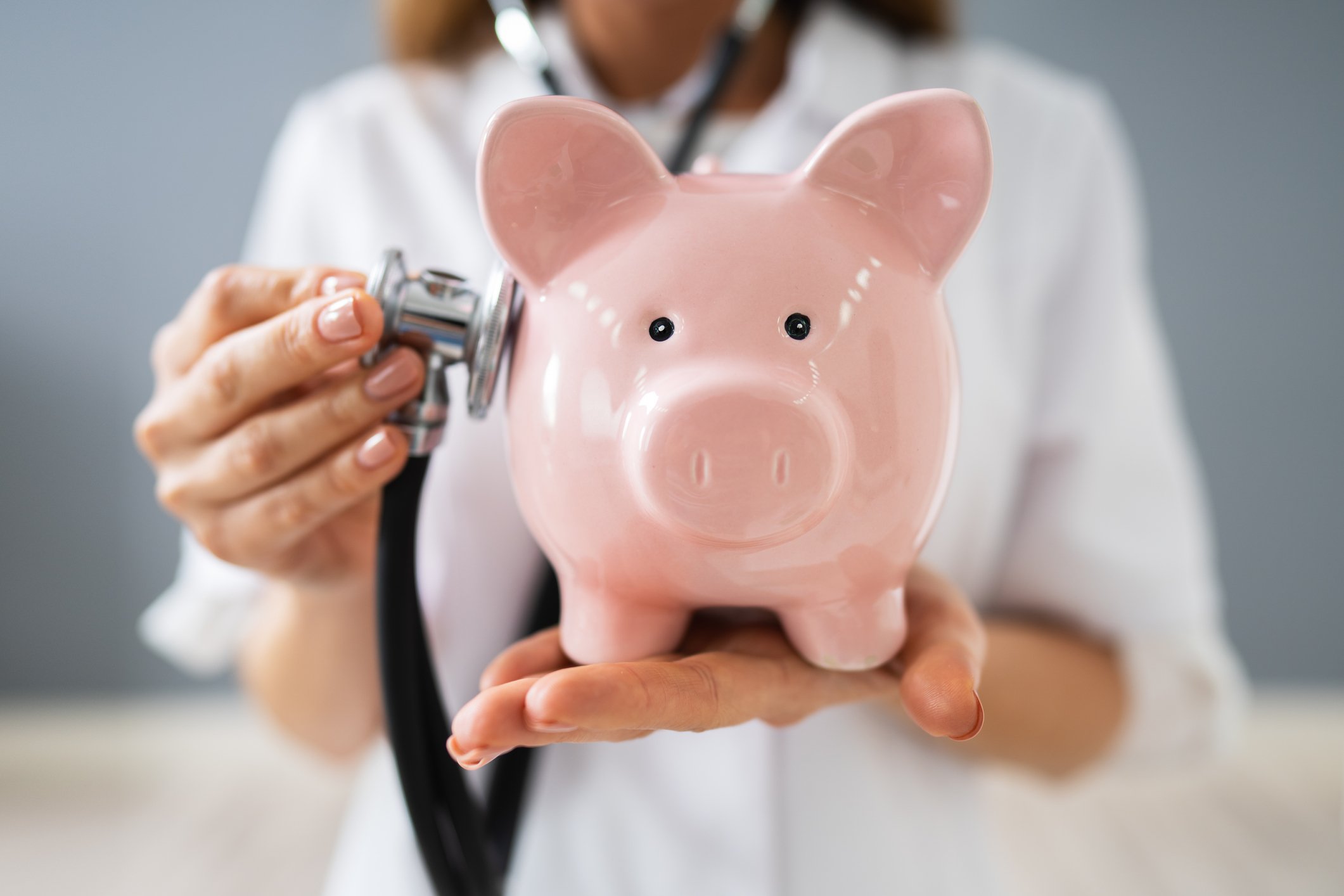 Woman in lab coat holding stethoscope to a piggy bank.