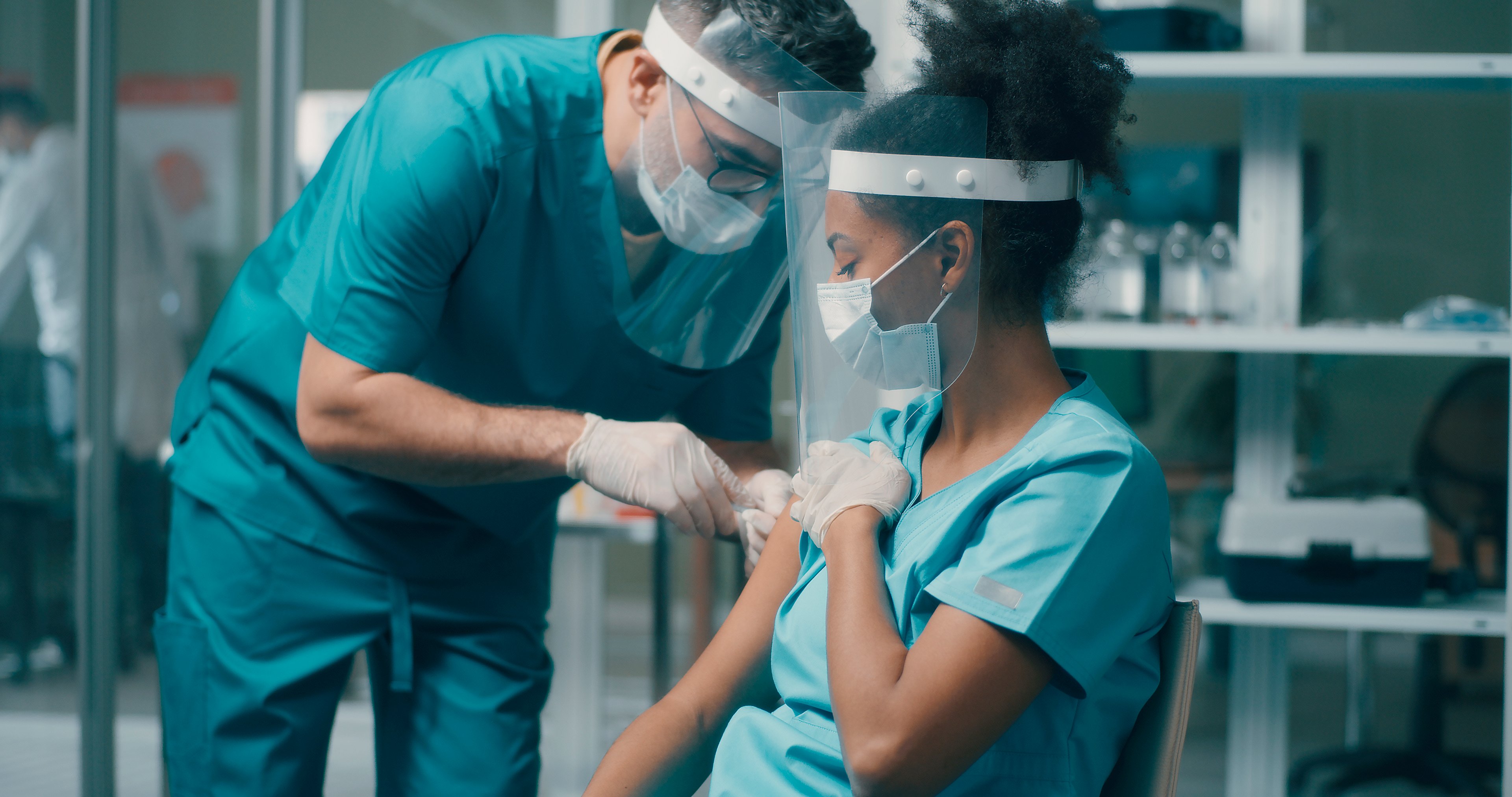 White male healthcare worker wearing a mask, glasses, green scrubs, and face shield administers a vaccine in the right arm of a seated African American woman wearing green scrubs, a mask, and a faceshield.