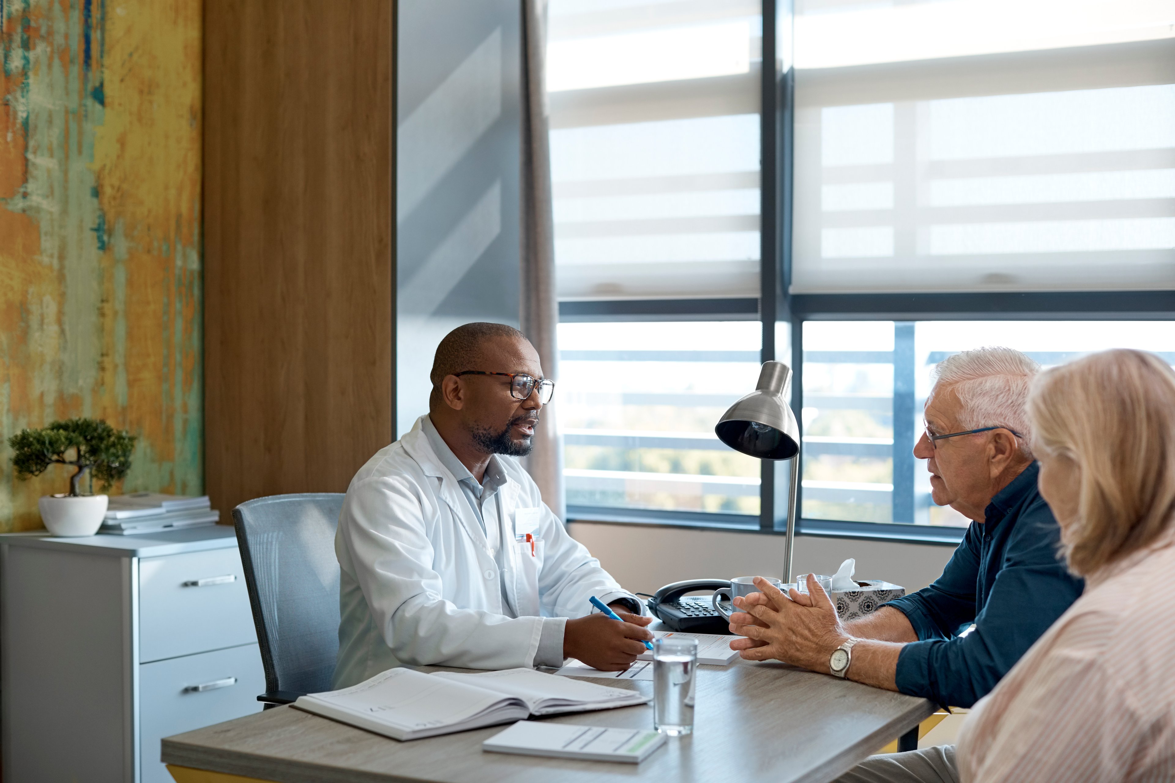 Black doctor wearing white physician's coat and glasses sits across a desk from a mature white male and female.