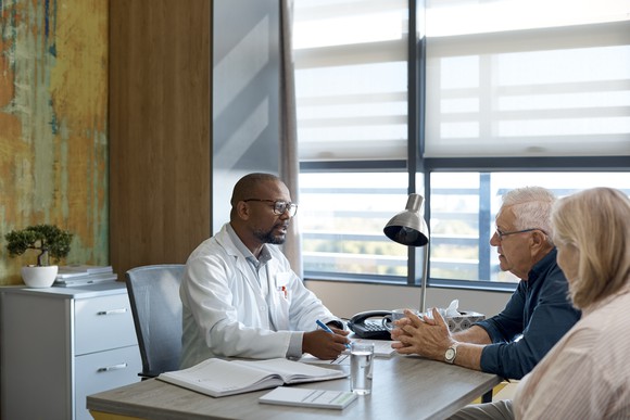 Black doctor wearing white physician's coat and glasses sits across a desk from a mature white male and female.