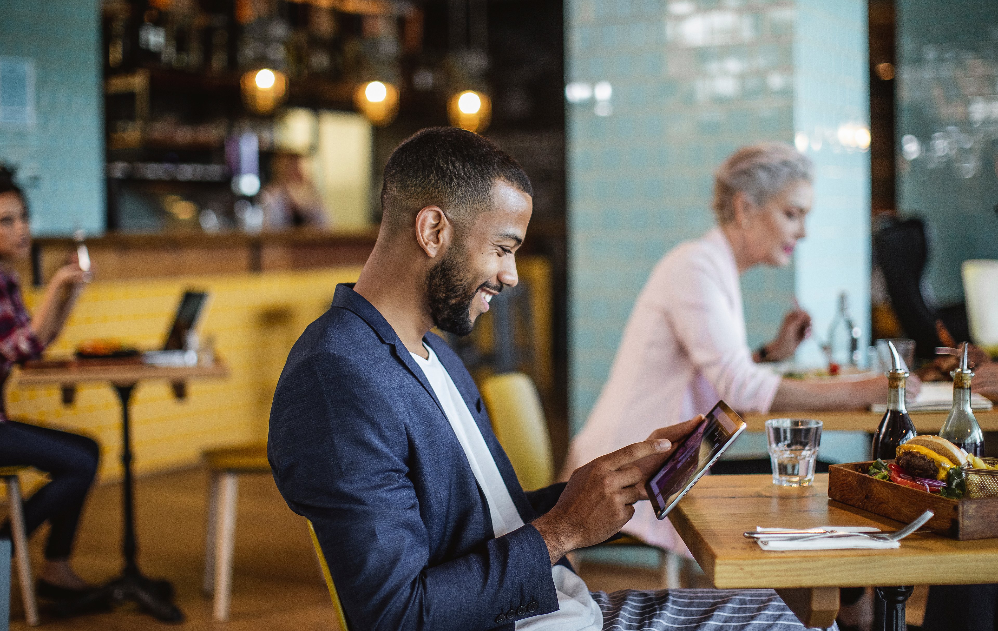 Young man wearing a sport jacket and white t-shirt sits at a cafe table, smiling while looking down at his tablet.