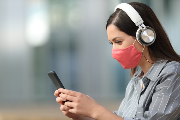 A woman wearing a red face mask and white headphones uses her smartphone.