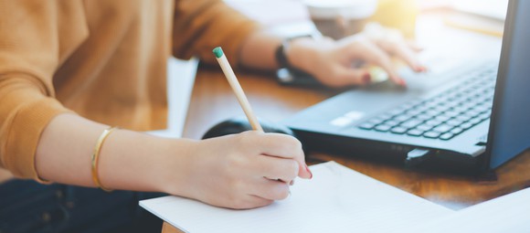 A woman takes notes with pencil and paper as she studies reports on her computer. 